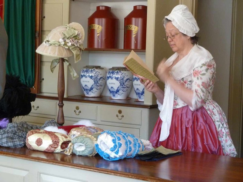 A photo of a woman in 18th century clothes reading a pamphlet in the Millinery Shop.