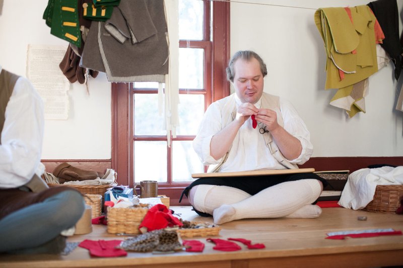 A photo of a man in 18th century clothing sitting cross-legged sewing.