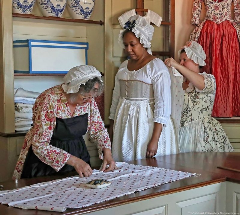 A photo of three women inside the Millinery Shop. One woman is folding fabric. The other two ladies are having a garment fitted.