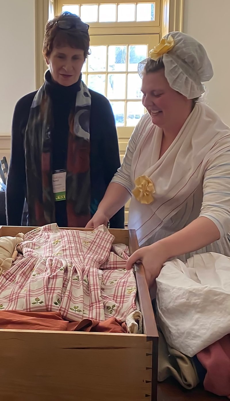 A photo of two women, one in modern day clothes and the other in 18th century clothes, looking at fabric in the Millinery Shop.